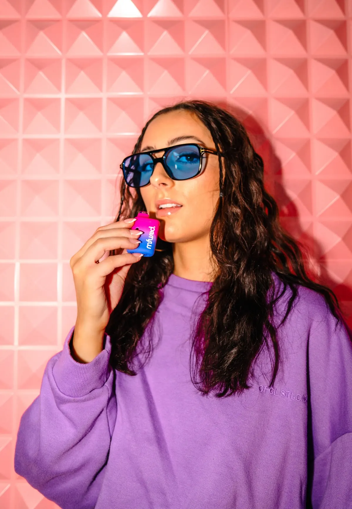 A woman in blue sunglasses holding a small pink product to her lips